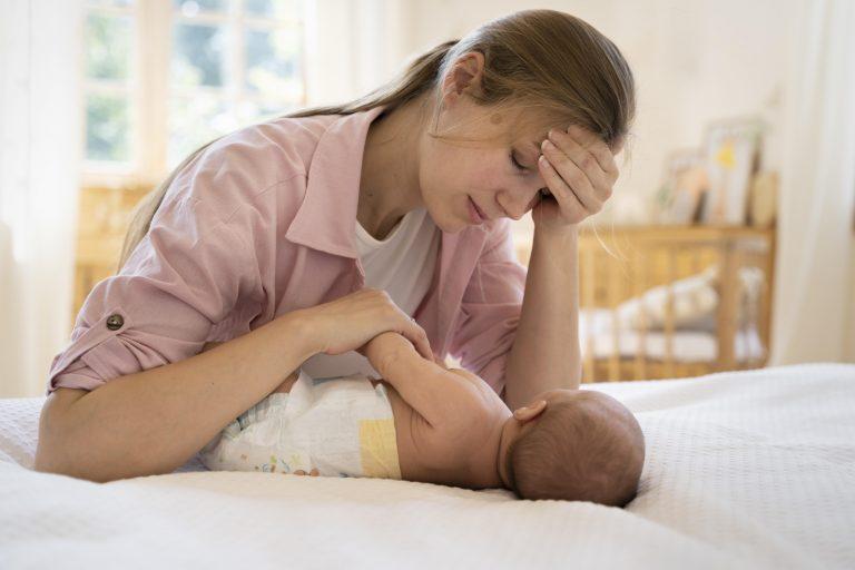 ermüdete Mutter im Wochenbett fasst sich an Stirn und kümmert sich um Baby