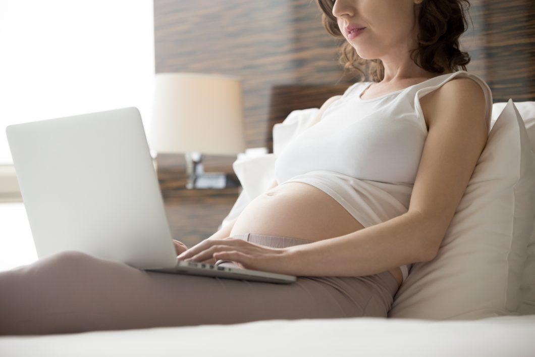 Schwangere Frau sitzt auf dem Sofa am Laptop und ist auf Hebammensuche