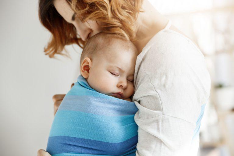 Mutter trägt schlafendes Baby mit blauem Tragetuch und neigt den Kopf nahe zu ihrem Kind.