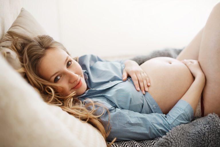Schwangere Frau liegt mit freiem Bauch auf dem Sofa und hält den Bauch in ihren Händen und lächelt in die Kamera.