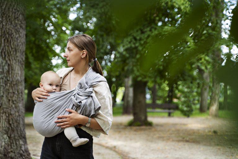 Mama trägt Baby auf dem Arm mithilfe eines grauen Tragetuchs.