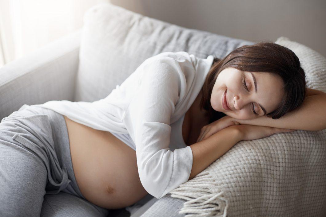 Schwangere liegt mit geschlossenen Augen und lächelnd auf dem Sofa.