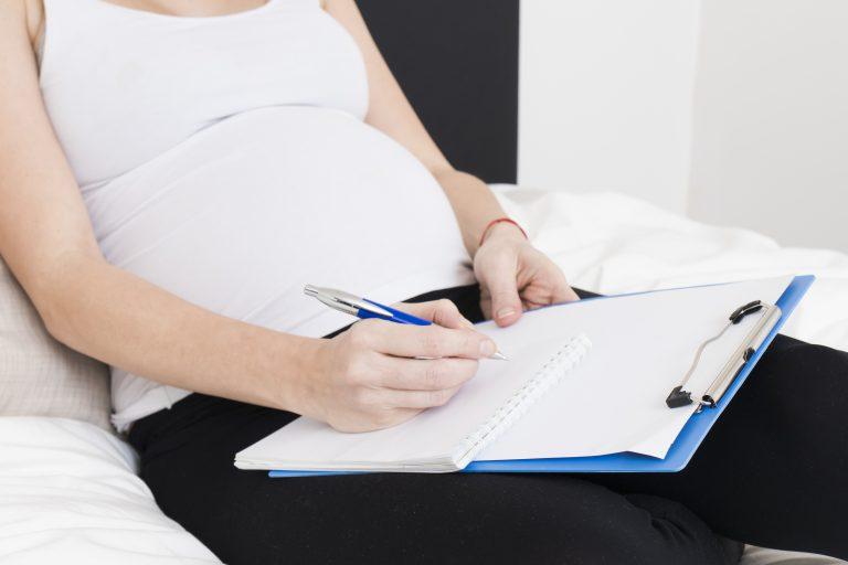 Schwangere Frau liegt auf dem Bett und macht sich auf einem Klemmbrett Notizen.