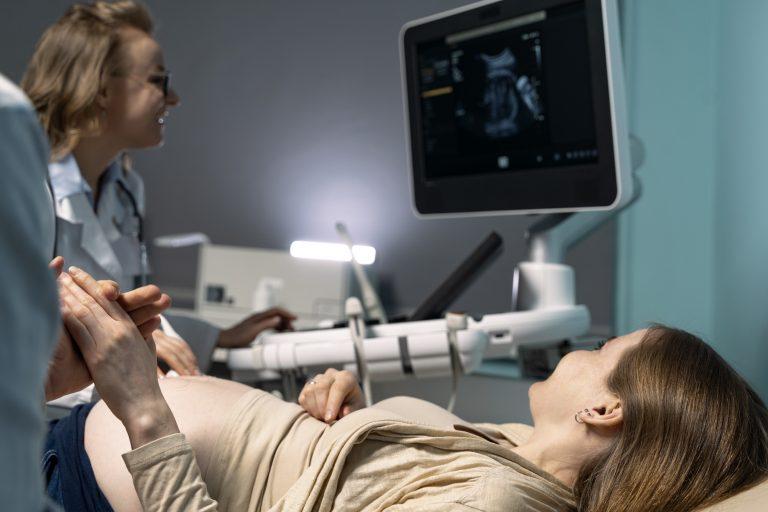 Frau hält Hand von Mann während Frauenärztin eine Ultraschalluntersuchung bei der schwangeren Frau durchführt.