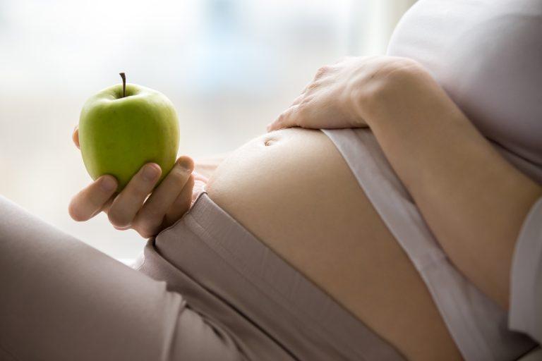 Schwangere Frau hält grünen Apfel vor ihren Babybauch.